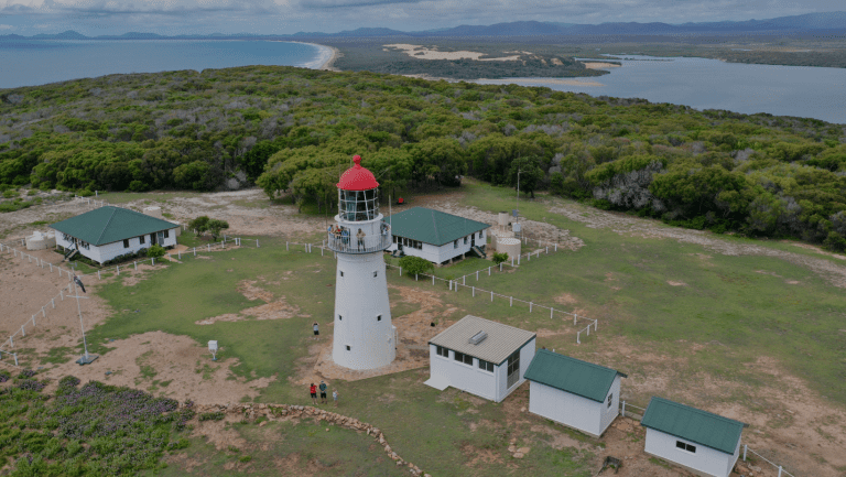 An aerial photo of the bustard headland light station. Including the lighthouse, people on the verandah and the ground. The two lighthouse keepers cottages. The engine room museum. Jenny Lind creek in the background as well as Bustard bay and mountain ranges.