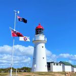 A photo of Bustard Head light house with blue skies in the background. On the left of the light house is a flag pole with three flags blowing in the wind.