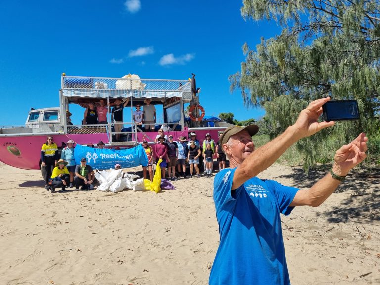Photo of a man taking a selfie of people standing with their bags of rubbish from the beach clean up day. LARC in the background and blue skies.