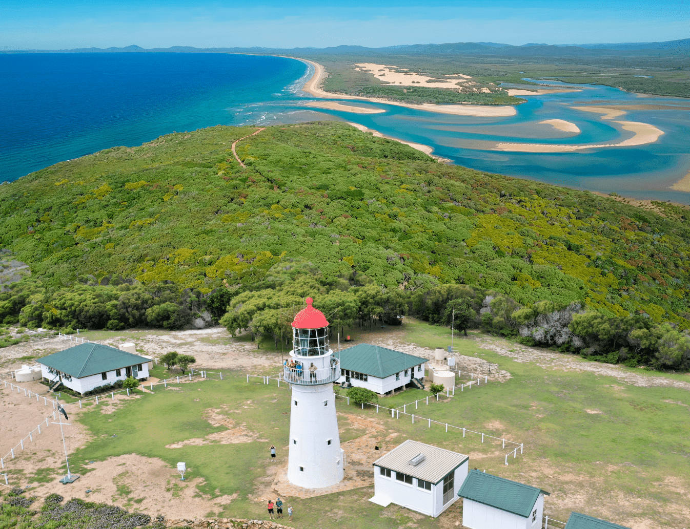 An aerial photo of the bustard head lighthouse station and its background of the native bushland of the headland, with the LARC road running through it. Behind that is crystal blue Jenny Lind creek with sand banks through it.